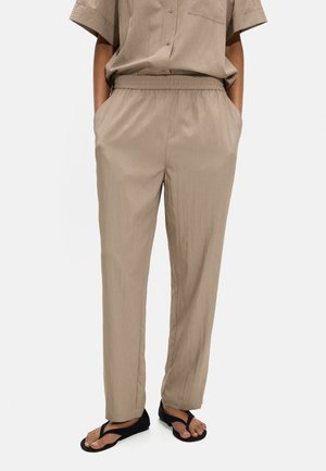 Osoba ubrana w beżowe spodnie z elastyczną talią, z rękami w kieszeniach i czarne sandały typu japonki, stojąca na jednolitym tle.