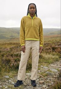 Žena s copánky, která má na sobě zelenou fleecovou bundu, béžové cargo kalhoty a černé turistické boty, stojí na kamenité stezce na travnatém svahu.