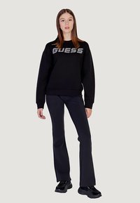 Must teksarihma dressipluus metallhalli "GUESS" logoga, mustade lehvikpükste ja mustade paksu tallaga tossudega.
