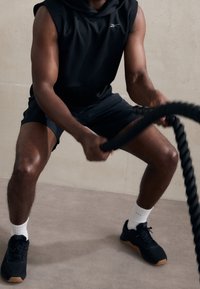 Svart ärmlös hoodie, svarta shorts och svarta träningsskor med gummisula. Håller i ett tjockt svart träningsrep. Slät golvbakgrund.