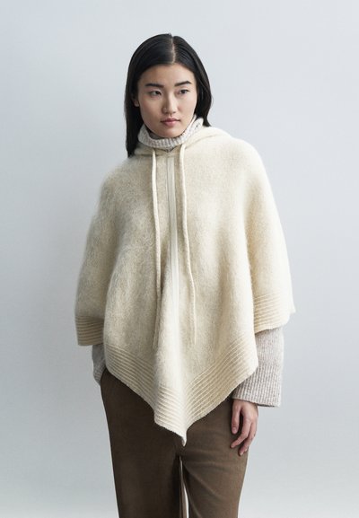Poncho en maille beige clair avec une finition texturée, doté d'une capuche, d'un cordon de serrage et d'un ourlet côtelé diagonal. Porté par-dessus un col roulé crème.