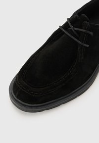 Schwarzer Wildlederschuh mit runder Spitze, strukturierten Oberflächen und sichtbaren Nähten. Die Schnürsenkel sind grau, und die Gummisohle hat ein geripptes Muster.