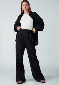 Črna prevelika srajca, bela rebrasta majica in široke črne hlače z vrvico. Gladek material, sproščen kroj in ravne sandale z zlatimi detajli.