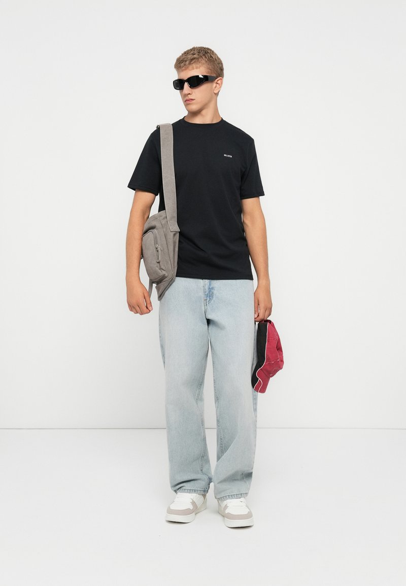 Młody mężczyzna noszący czarną koszulkę, jasnoniebieskie dżinsy, białe trampki, czarne okulary przeciwsłoneczne, noszący szary plecak i trzymający czerwoną czapkę bejsbolową.