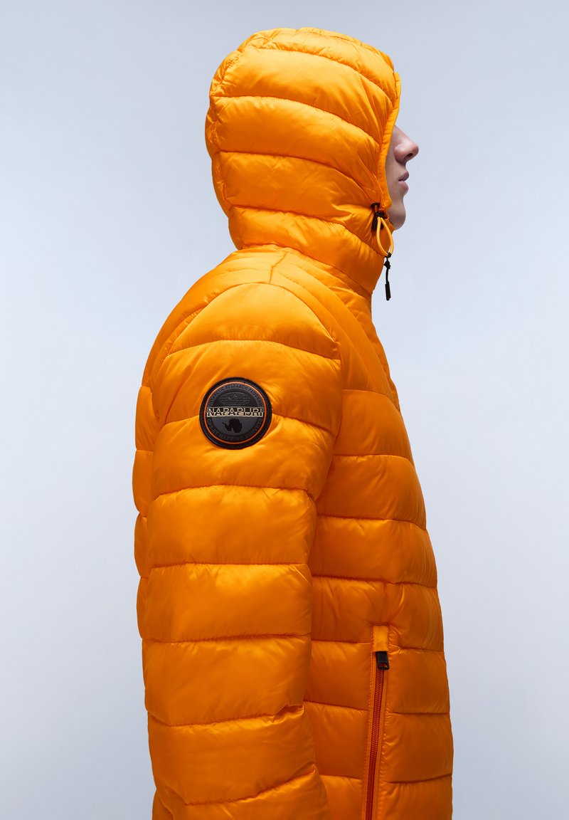 Chaqueta naranja acolchada con capucha, que cuenta con un bolsillo frontal con cremallera y un parche de logo circular en la manga izquierda. Textura suave.