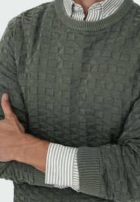 Zeleni teksturirani džemper s geometrijskim uzorkom, s rebrastim manžetama i izrezom, složen preko bijele i zelene prugaste košulje.