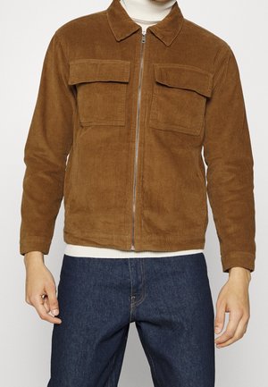 Mann trägt eine braune Cord-Zip-up-Jacke mit Klappentaschen über einem weißen Rollkragenpullover und dunkelblauen Jeans, steht vor einem einfarbigen Hintergrund.