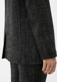 Blazer nero texture realizzato in tessuto intrecciato, con un taglio dritto e una piccola apertura laterale, esposto sulla mano di un modello.