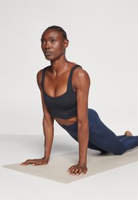 Μαύρο αθλητικό σουτιέν και σκούρο μπλε κολάν σε ένα άτομο που βρίσκεται σε θέση σανίδας πάνω σε ένα μπεζ υλικό γιόγκα, με ορατή μυϊκή διαφοροποίηση και εστίαση.