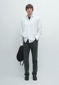 Weißes Hemd mit Knöpfen, dunkelgraue Jeans, schwarze Schuhe. Modell hält eine braune Jacke. Schlichter Hintergrund. Lässiger Schnitt, glattes Material, schlichtes Design.