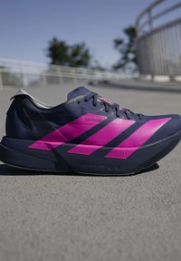 Chaussure de course athlétique bleu foncé et rose vif sur le trottoir avec une rambarde floue et des arbres en arrière-plan.