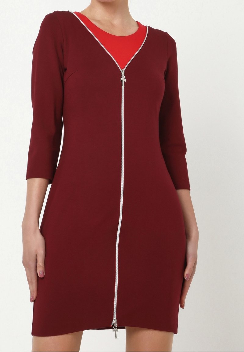 Femme portant une robe bordeaux ajustée avec une fermeture éclair argentée sur toute la longueur à l'avant et des manches trois-quarts, par-dessus un haut intérieur rouge.