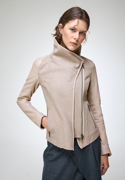 Femme portant une veste beige asymétrique zippée avec des manches en cuir et un pantalon gris foncé, debout devant un fond clair uni.