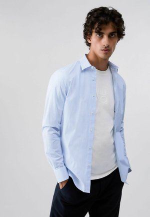 Jeune homme aux cheveux bouclés portant une chemise bleu clair boutonnée sur un t-shirt blanc et un pantalon sombre, debout les mains dans les poches.