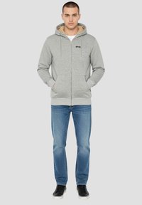 Sweat à capuche gris zippé avec une doublure intérieure douce, deux poches avant et une capuche avec cordon de serrage, associé à un jean bleu et des baskets noires.