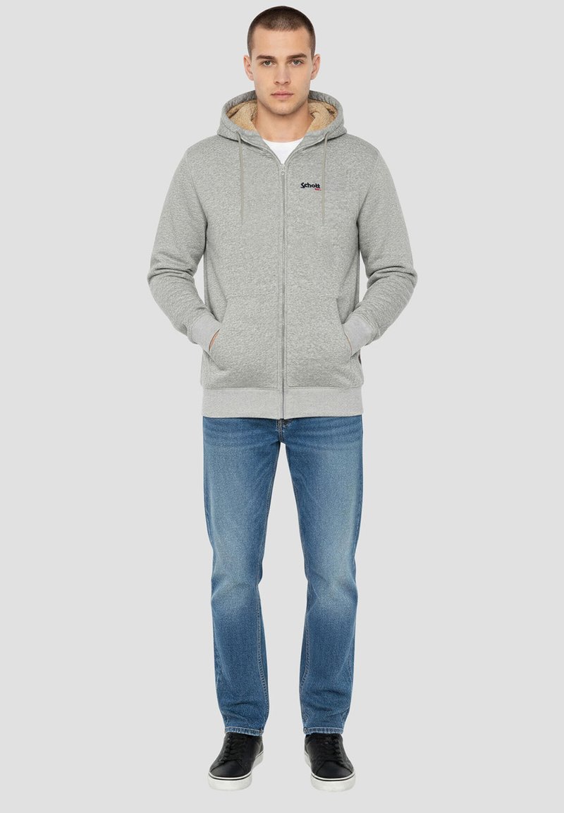 Sweat à capuche gris zippé avec une doublure intérieure douce, deux poches avant et une capuche avec cordon de serrage, associé à un jean bleu et des baskets noires.