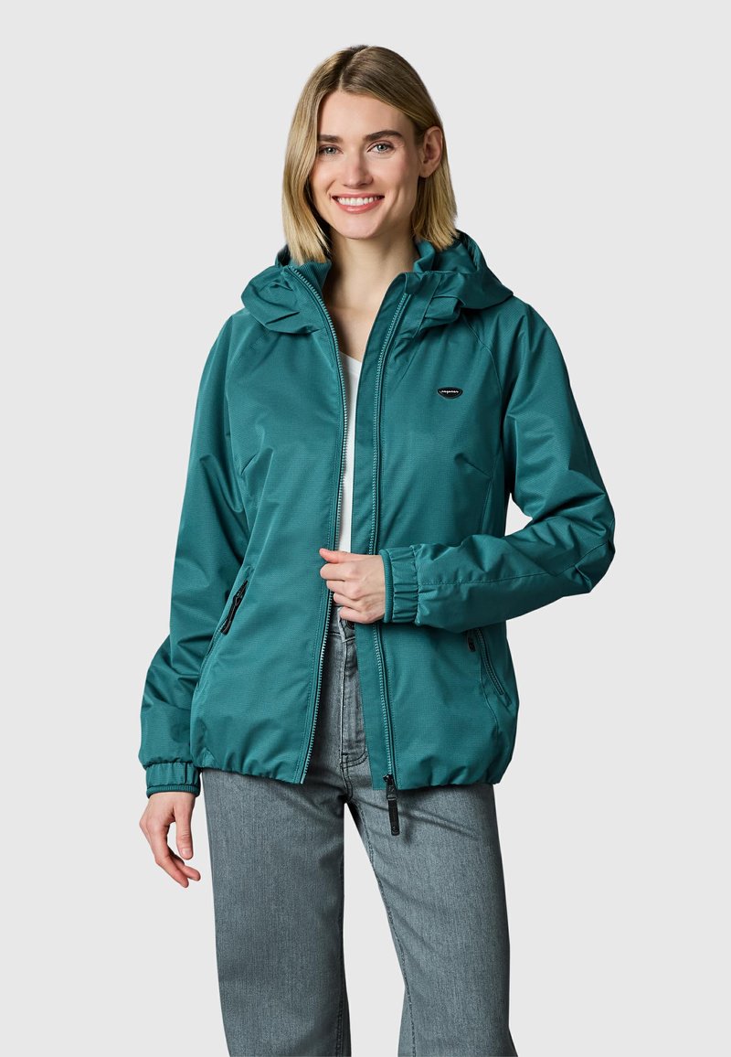 Lachende vrouw die een turquoise capuchonjack met rits en lichtgrijze spijkerbroek draagt, staand tegen een effen lichtgrijze achtergrond.