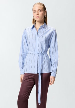 Donna con camicia a righe azzurro chiaro con bottoni e dettagli in perle, cintura annodata in vita, abbinata a pantaloni bordeaux scuro.