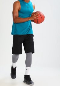 Persona che indossa una maglietta senza maniche color turchese, pantaloni corti neri, leggings grigi e sneakers nere, mentre tiene un pallone da basket con entrambe le mani.