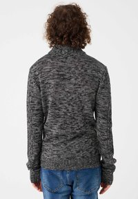 Suéter gris de punto con cuello alto, que presenta un patrón texturizado. Mangas de corte holgado y dobladillo acanalado. Llevado con jeans de mezclilla azul.