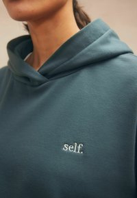 Kapišonas džemperis muted teal spalvos audinyje, su maža balta išsiuvinėta detale "self." ant krūtinės. Minkštas tekstūros ir laisvas tinkantis.