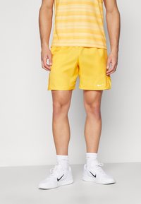 Miestenské žlté športové šortky so bočnými rozparky, vyrobené z ľahkého materiálu, s elastickým pásom a malým bielym logom Nike. Skombinované s bielymi topánkami.
