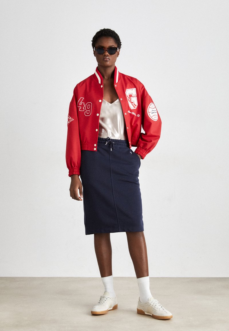 Czerwona kurtka typu varsity z białymi akcentami, satynowy top na ramiączkach, granatowa spódnica ołówkowa i białe sneakersy. Modelka prezentuje zgrabny, miejski strój z minimalną teksturą.