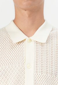 Pleteni polo majica v off-white barvi s teksturiranim, odprtim tkanjem. Ima klasičen ovratnik in zapiranje na gumb.