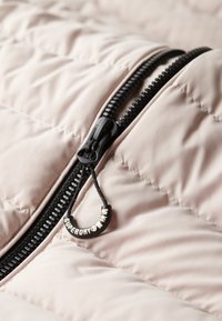 Giacca imbottita in morbido tessuto rosa pallido con zip nera e linguetta testurizzata. La zip presenta dettagli brandizzati per un tocco in più.