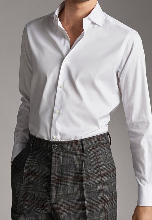 Man in een witte lange mouw knoop-blouse, gestoken in donkere grijze geruite broek met handen in de zakken.