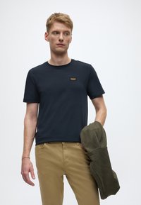 T-shirt en coton bleu marine à manches courtes et col rond. Présente un petit patch logo beige sur la poitrine. Veste vert olive drapée sur le bras.