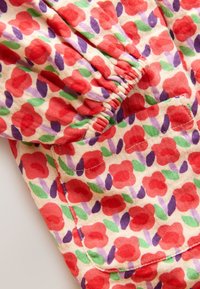 Blomstret stof med en lys cremefarvet baggrund, der har røde blomster, grønne blade og lilla detaljer, med elastik gathered ved halsudskæringen og lommen.
