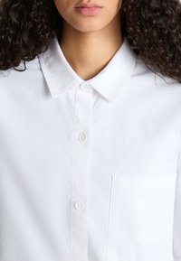 Camisa blanca de botones con cuello, con una textura de tela suave, un pequeño bolsillo en el pecho y cinco botones en la parte delantera.