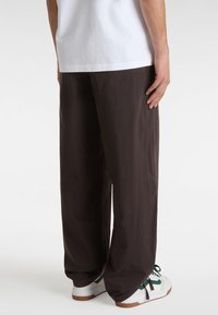 Brown, loose-fitting trousers with a textured finish. Features side pockets and a relaxed silhouette, paired with white and green sneakers.