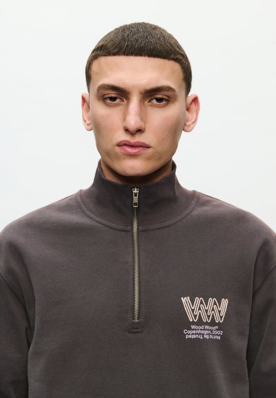 Tmavošedá mikina na zips s vysokým golierom, s bielym vyšívaným logom na hrudi a hladkým, textúrovaným materiálom.