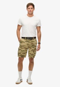 Camo cargo shorts i khaki med flere lommer, parret med en tætsiddende hvid t-shirt. Accessories inkluderer et sort bælte og hvide sportsokker.