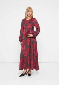 Długo-sleeve, kwiecista sukienka maxi w ciemnym burgundzie z czerwonymi kwiatami. Modelka trzyma bordową kopertówkę i ma na sobie czarne sandały na obcasie.