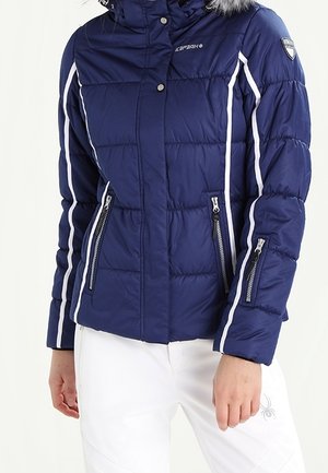 Veste matelassée bleu marine avec des rayures blanches, poches zippées à l'avant et sur la manche, portée avec un pantalon blanc avec un petit logo araignée.