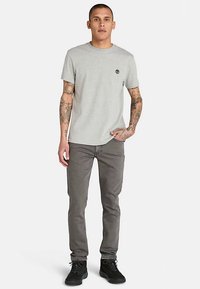 Jasnoszary bawełniany t-shirt z okrągłym dekoltem i krótkimi rękawami, w połączeniu z ciemnoszarymi dżinsami i czarnymi butami. Na piersi znajduje się małe logo.