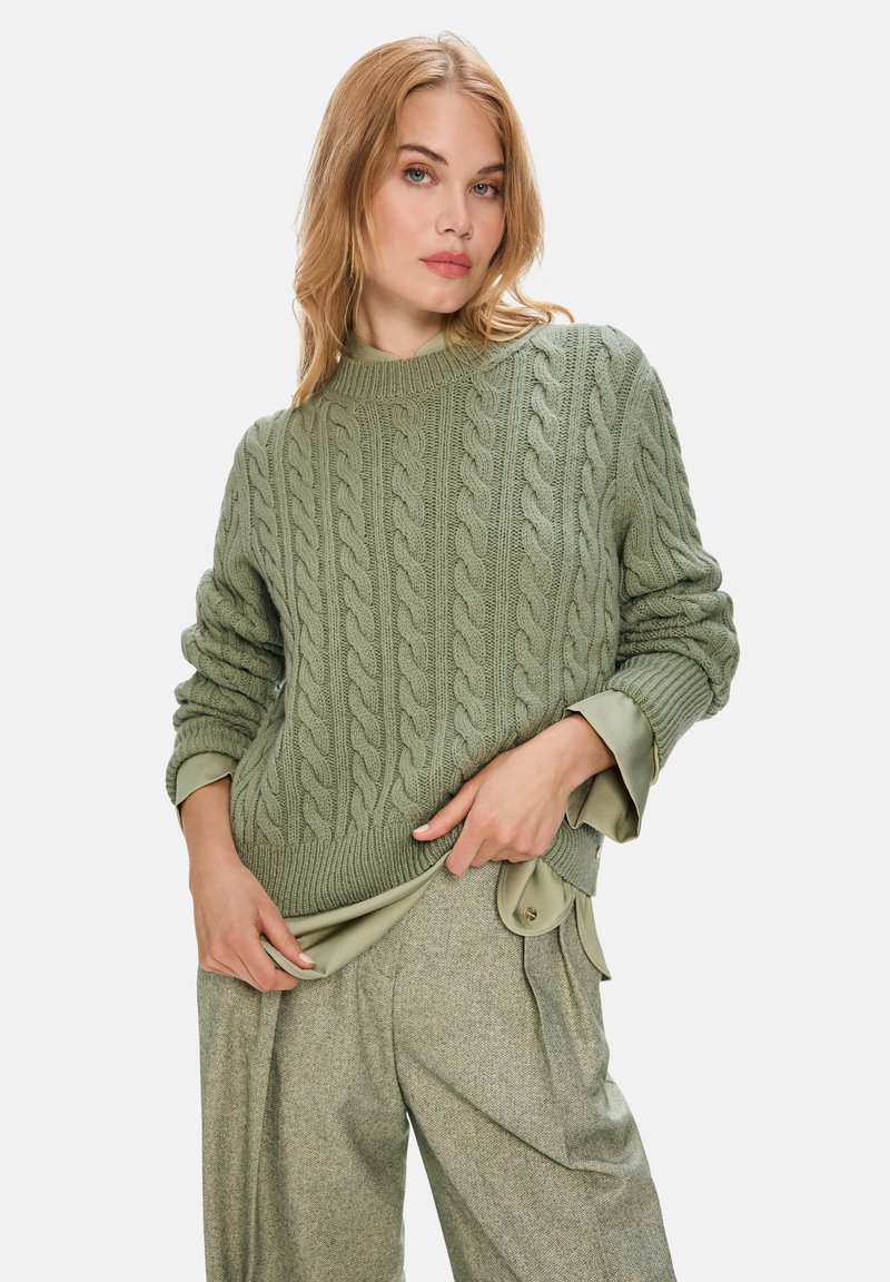 Grüner Kabelstrickpullover mit hohem Kragen und langen Ärmeln, kombiniert mit einem passenden strukturierten grünen Outfit. Weiche, voluminöse Stricktextur.