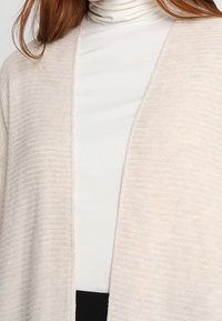 Ljusbeige kardiagan med öppen framsida, ribbat mönster och långa ärmar, lagd över en vit polotröja. Ingen knäppning eller försköningar.
