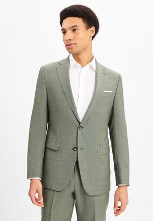 Jonge man in een lichtgroed tweeknops pak met een wit overhemd, staand en iets naar de zijkant kijkend tegen een witte achtergrond.