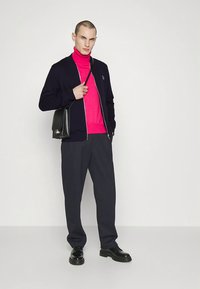 Marineblå cardigan med glidelås over en knallrosa turtleneck, kombinert med mørke bukser og svarte sko, tilbehør med en svart skulderveske.