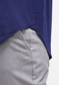 Granatowa bawełniana bluzka z zaokrąglonym dołem, zestawiona z jasnoszarymi, teksturowanymi spodniami z boczną kieszenią. Zbliżenie.