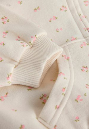 Kreemikas pusa soonikkoes varrukapitsidega, millel on väiksed roosad lillemustrid ja rohelised lehed üle kogu kanga, rõhutades pehmet tekstuuri.