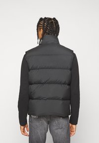 Gilet imbottito nero con colletto alto, caratterizzato da una trapuntatura orizzontale, imbottitura leggera e una finitura elegante. Indossato sopra una camicia a maniche lunghe.