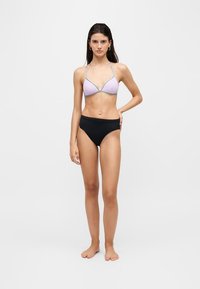Femme debout pieds nus, portant un haut de bikini violet clair avec un bord argenté et un bas de bikini noir taille haute sur fond blanc uni.