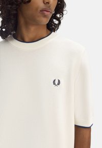Polo a maniche corte di colore crema, lavorato a maglia con un motivo testurizzato e un bordo blu navy nel colletto e nelle maniche. Presenta un piccolo logo sul petto.