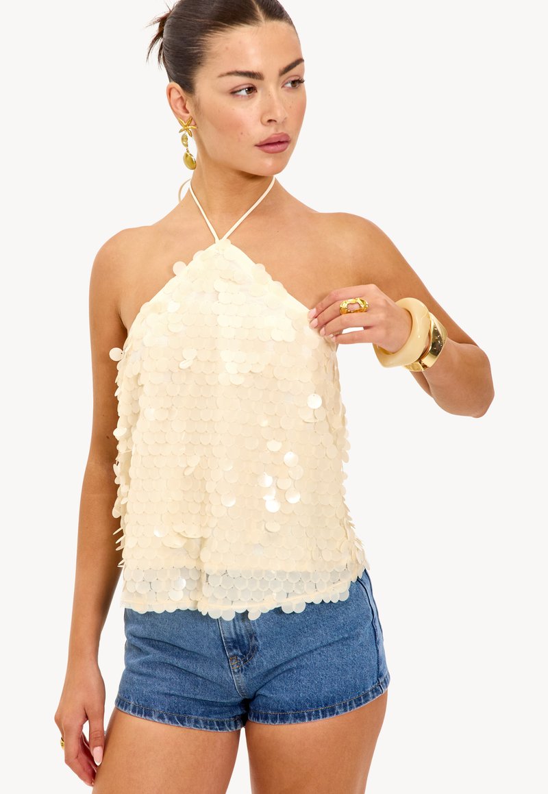Femme portant un haut halter sans manches en sequins blancs, un short en denim, et des boucles d'oreilles, bagues et bracelets en ton doré, regardant sur le côté.