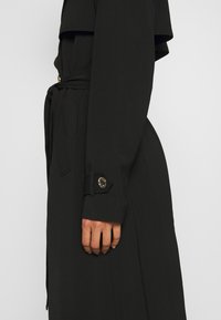Vue de côté d'une personne portant un trench-coat noir à manches longues avec une ceinture nouée à la taille et un détail de poignet boutonné.